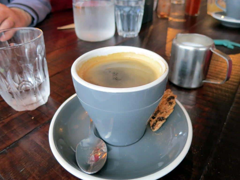 A flat white coffee in a white ceramic cup next to almond biscotti on a wooden table.