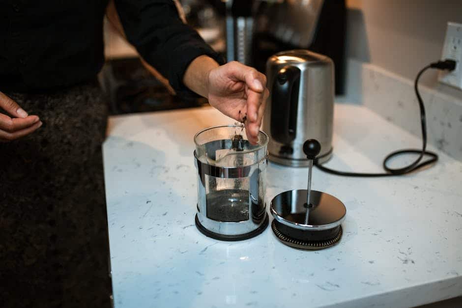 A person's hands pressing down the plunger of a French press and then pouring the brewed coffee into a cup, demonstrating proper technique